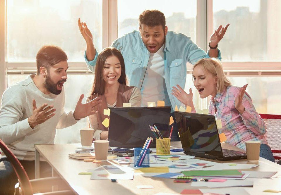 Spaß im Team sorgt für einen angenehmen Alltag im Büro. Ein Team aus zwei jungen Männern und zwei jungen Frauen sitzt an einem großen Tisch um einen Laptop herum, sie lachen und machen überraschte Gesichter und Gesten.