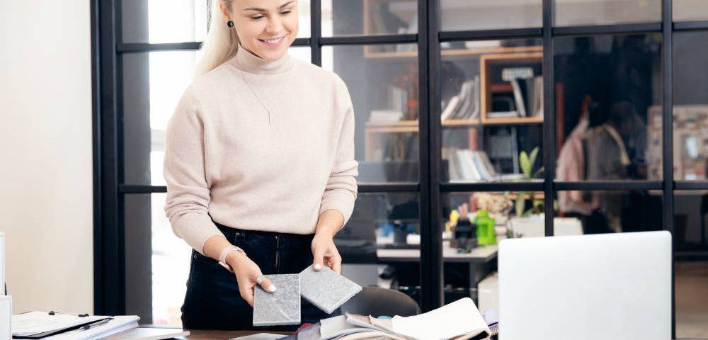 Eine junge Frau mit blonden, hinten zusammengenommenen Haaren, schwarzer Hose und hellem Pulli steht in einem modern gestalteten Büro vor einem Laptop auf dem Schreibtisch und plant mit Farbkarten und Stoffproben die Büroeinrichtung.