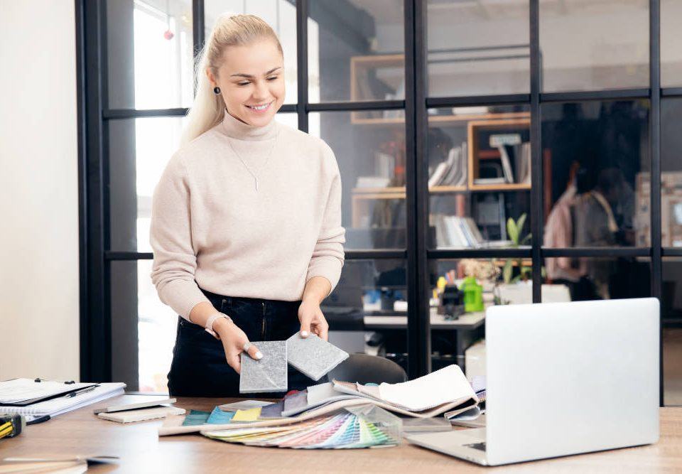 Eine junge Frau mit blonden, hinten zusammengenommenen Haaren, schwarzer Hose und hellem Pulli steht in einem modern gestalteten Büro vor einem Laptop auf dem Schreibtisch und plant mit Farbkarten und Stoffproben die Büroeinrichtung.