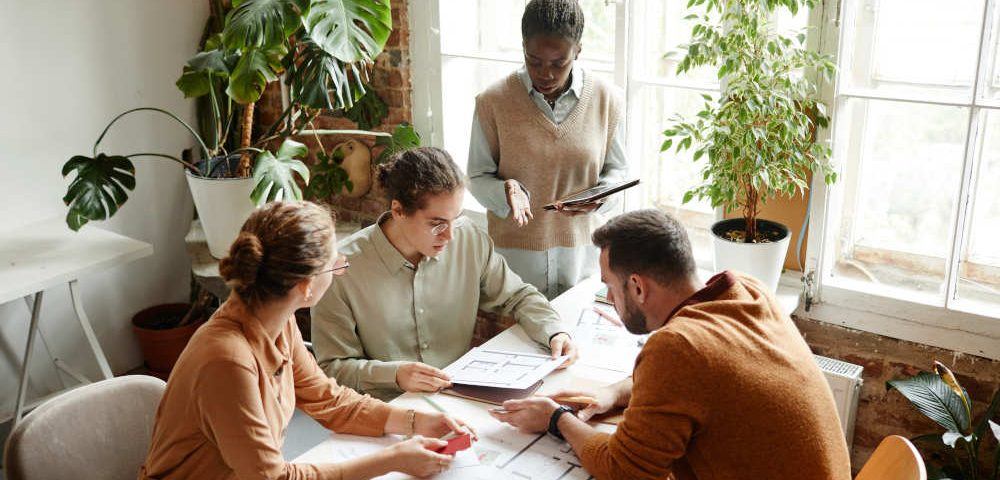 Ein vierköpfiges Team sitzt einem modernen Büro zusammen an einem Tisch und diskutiert angeregt, im Hintergrund sind grüne Büropflanzen