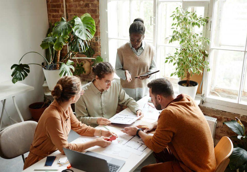 Ein vierköpfiges Team sitzt einem modernen Büro zusammen an einem Tisch und diskutiert angeregt, im Hintergrund sind grüne Büropflanzen