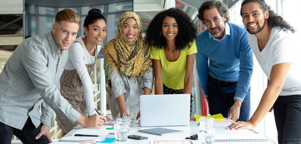 Diversity am Arbeitsplatz: Ein sechsköpfiges Team, drei Frauen und drei Männer unterschiedlichen Alters und Herkunft stehen vor einem Schreibtisch in einem Büro und lächeln in die Kamera