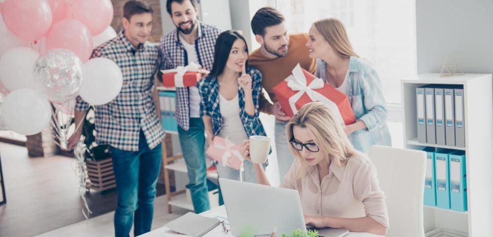 Geschenke fürs Büro: Fünf Kollegen schleichen sich mit Luftballons und verpackten Geschenken an den Schreibtisch einer Kollegin heran, um sie zu überraschen