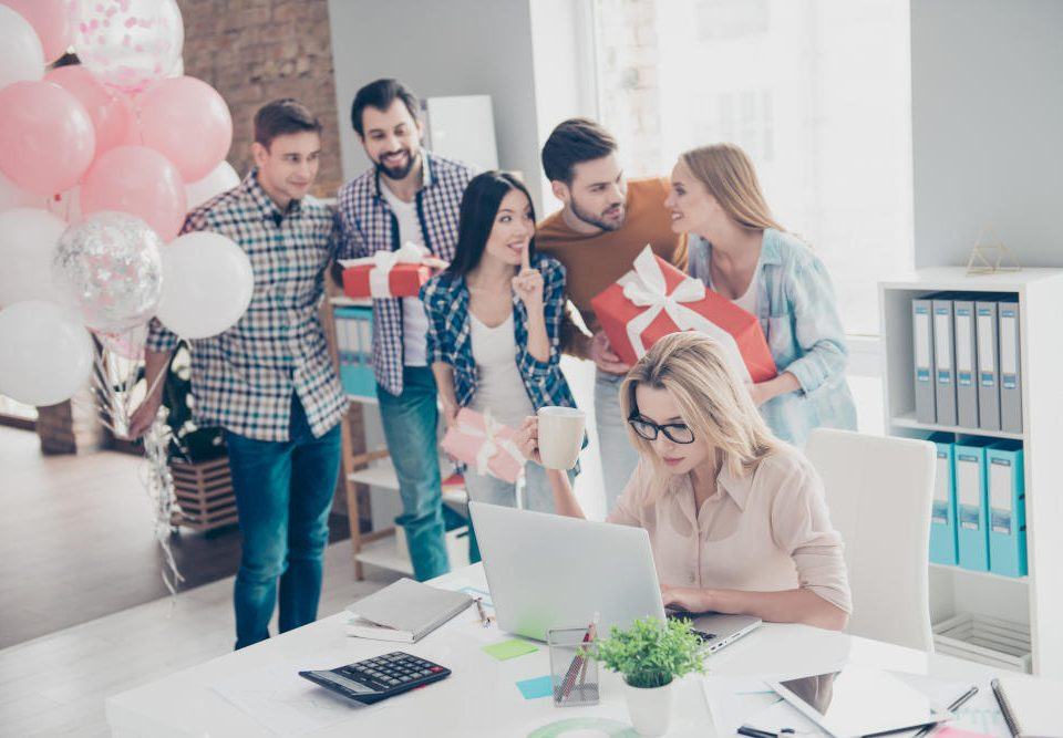 Geschenke fürs Büro: Fünf Kollegen schleichen sich mit Luftballons und verpackten Geschenken an den Schreibtisch einer Kollegin heran, um sie zu überraschen