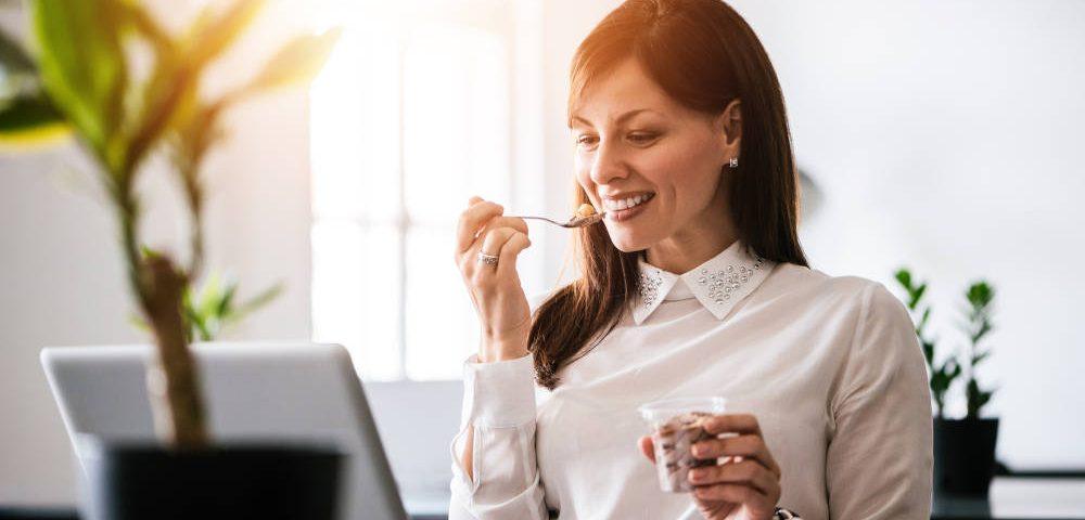 Eine junge, dunkelhaarige Frau sitzt bei Sommer im Büro. Sie schaut lächelnd auf einen Laptop, der auf dem Schreibtisch vor ihr steht, und löffelt gegen die Hitze im Büro ein Eis. Durch das Fenster scheint die Sonne herein, im Vordergrund steht eine grüne Topfpflanze auf dem Schreibtisch.