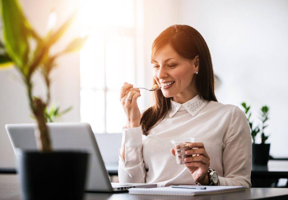 Eine junge, dunkelhaarige Frau sitzt bei Sommer im Büro. Sie schaut lächelnd auf einen Laptop, der auf dem Schreibtisch vor ihr steht, und löffelt gegen die Hitze im Büro ein Eis. Durch das Fenster scheint die Sonne herein, im Vordergrund steht eine grüne Topfpflanze auf dem Schreibtisch.