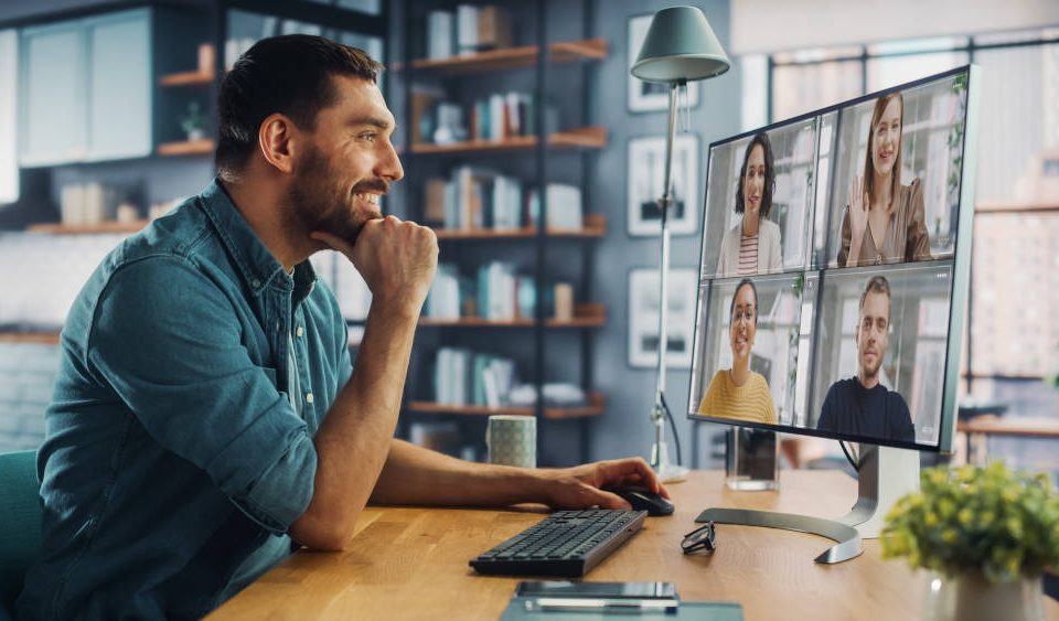 Home Office Vor- und Nachteile: Ein junger Mann mit Bart sitzt an seinem Schreibtisch zu Hause und unterhält sich per Videokonferenz mit vier Kollegen.