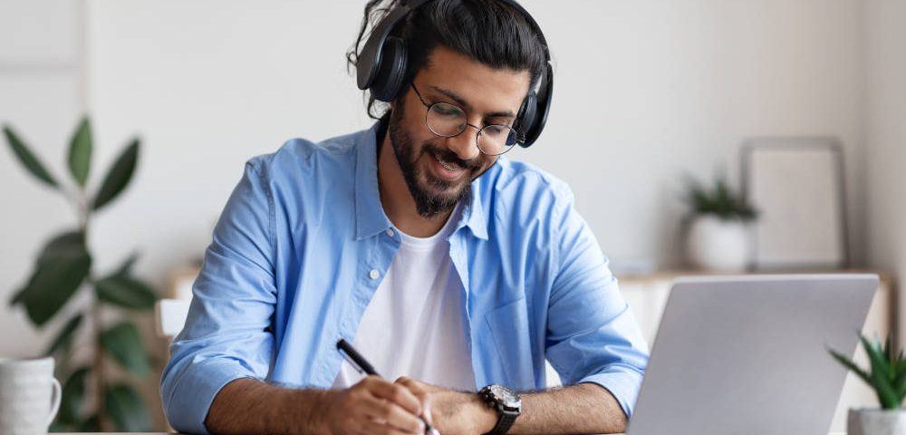 Ein junger Mann mit lange, hinten zusammengebundenen schwarzen Haaren und Bart sitzt in einem hellen Raum vor seinem Laptop. Er hört mit großen Kopfhörern Musik am Arbeitsplatz, während der sich handschriftliche Notizen macht.