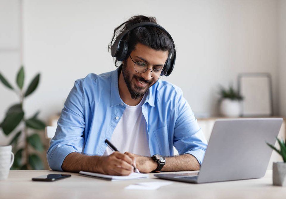 Ein junger Mann mit lange, hinten zusammengebundenen schwarzen Haaren und Bart sitzt in einem hellen Raum vor seinem Laptop. Er hört mit großen Kopfhörern Musik am Arbeitsplatz, während der sich handschriftliche Notizen macht.