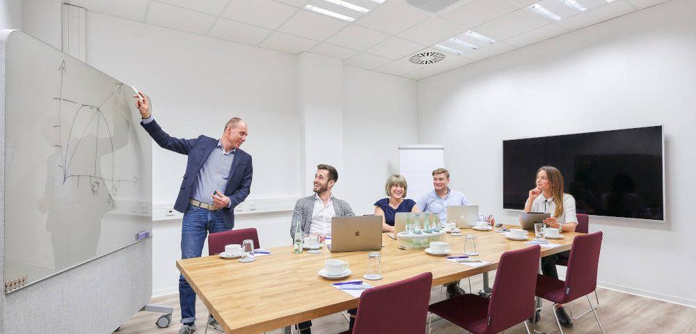 Ein etwas älterer Mann steht in einem Meetingraum am Whiteboard und hält eine Präsentation. Er zeichnet ein Diagram an und wendet sich zu seinen Zuhörern, zwei jüngeren Männern und zwei Frauen, die um einen viereckingen Besprechungstisch herum sitzen und aufmerksam zuhören.