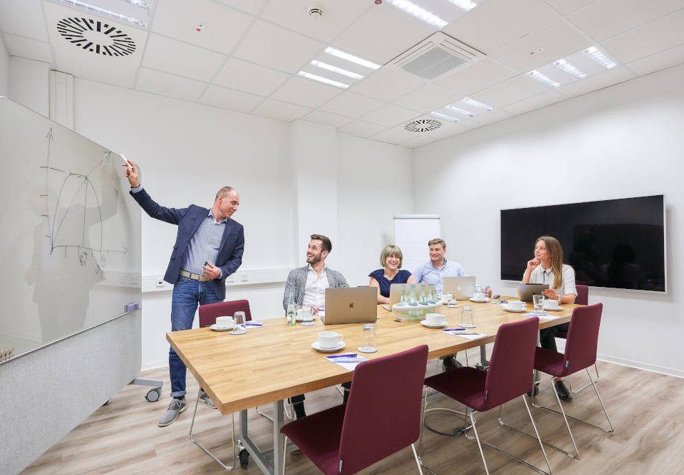 Ein etwas älterer Mann steht in einem Meetingraum am Whiteboard und hält eine Präsentation. Er zeichnet ein Diagram an und wendet sich zu seinen Zuhörern, zwei jüngeren Männern und zwei Frauen, die um einen viereckingen Besprechungstisch herum sitzen und aufmerksam zuhören.