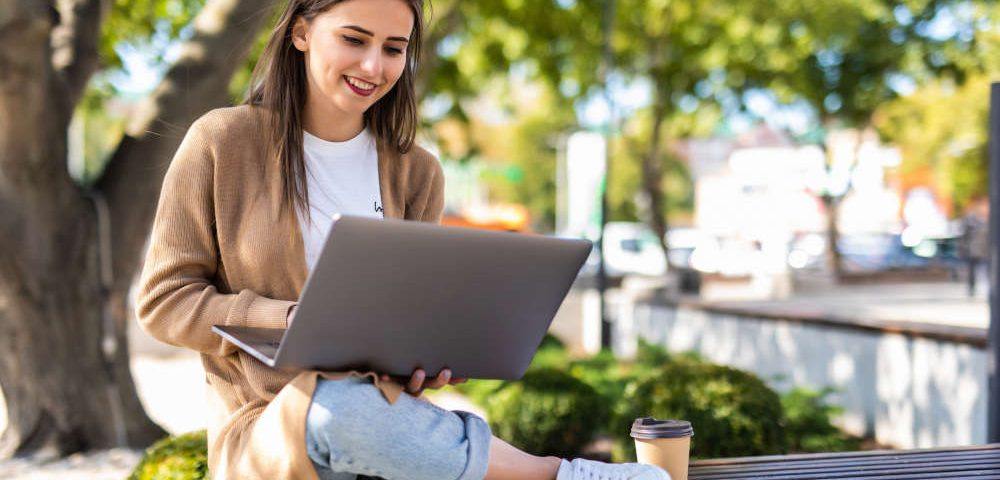 Virtuelle Geschäftsadresse mieten und überall arbeiten: Eine junge Frau in heller Jeans und Cardigan sitzt in der Sonne auf einer Bank unter einem Baum, neben ihr steht ein Coffee-to-go-Becher. Sie lächelt und tippt auf einem Laptop, den sie auf den Knien hält.