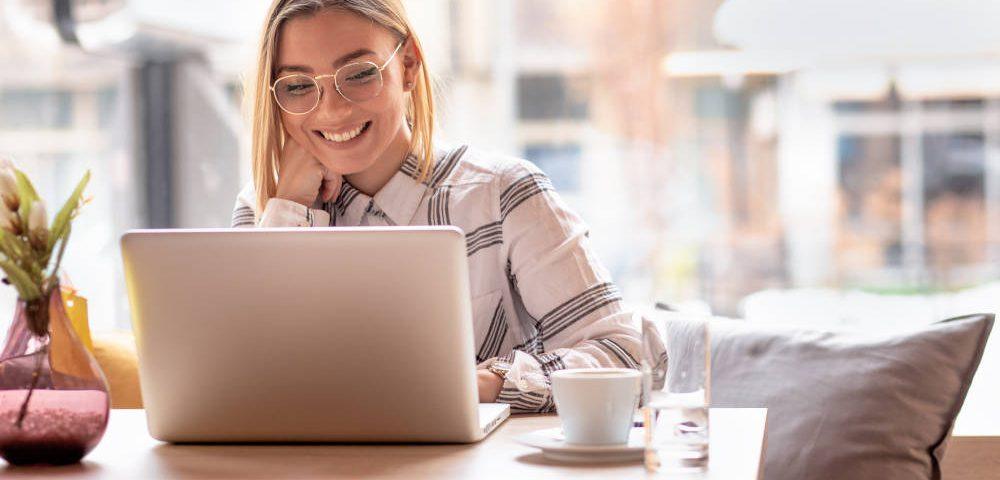 Junge Frau sitzt mit Kaffeetasse lachend am Laptop, genießt die Vorteile der Selbstständigkeit