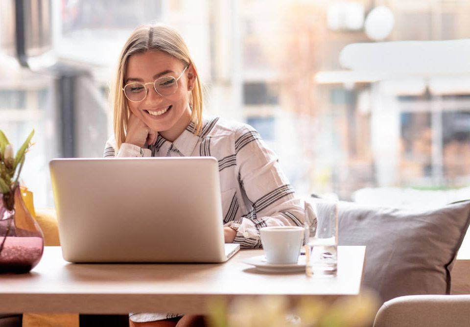 Junge Frau sitzt mit Kaffeetasse lachend am Laptop, genießt die Vorteile der Selbstständigkeit
