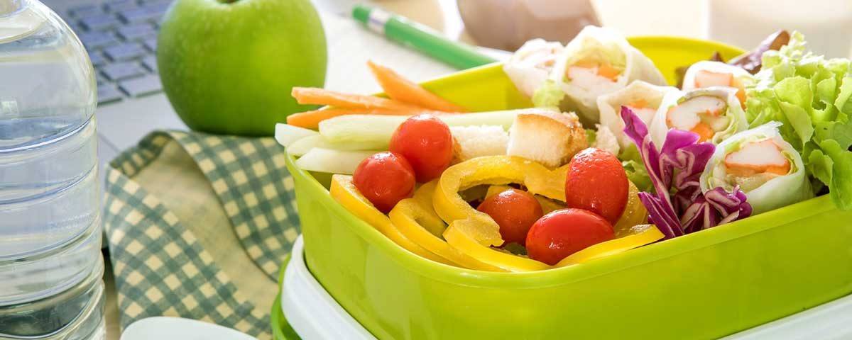 Eine grüne Tupperbox steht auf einem Schreibtisch, gefüllt mit frischem Obst und Gemüse. Daneben liegt ein grüner Apfel. So geht Gesunde Ernährung im Büro!