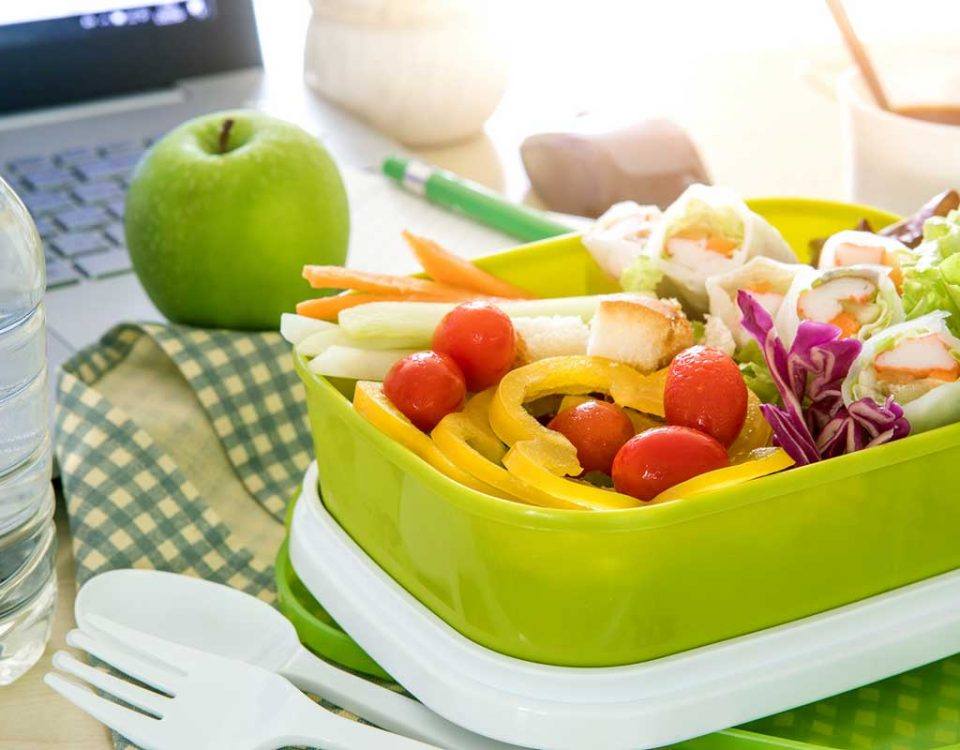 Eine grüne Tupperbox steht auf einem Schreibtisch, gefüllt mit frischem Obst und Gemüse. Daneben liegt ein grüner Apfel. So geht Gesunde Ernährung im Büro!