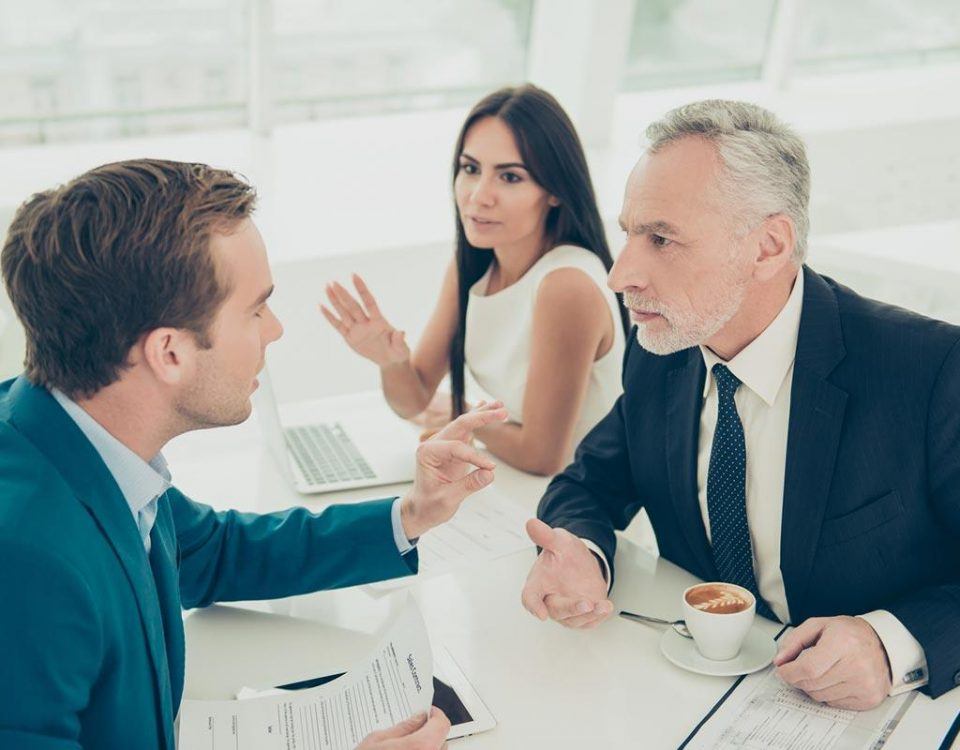 Kommunikation im Team: Eine Frau, ein älterer und ein jüngerer Mann sitzen in einem Meetingraum um einen Tisch herum. Beide Männer tragen einen Anzug. Die drei sind in eine angeregte Diskussion verwickelt.