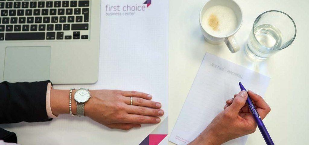 Auf einem weißen Schreibtisch steht ein silberner Laptop, daneben eine Kaffeetasse und ein Wasserglas sowie ein Schreibblock mit dem Logo der First Choice Business Center. Eine Person, von der man nur die Arme und Hände sieht, macht sich Notizen zum Thema Zeitmanagement auf dem Block.