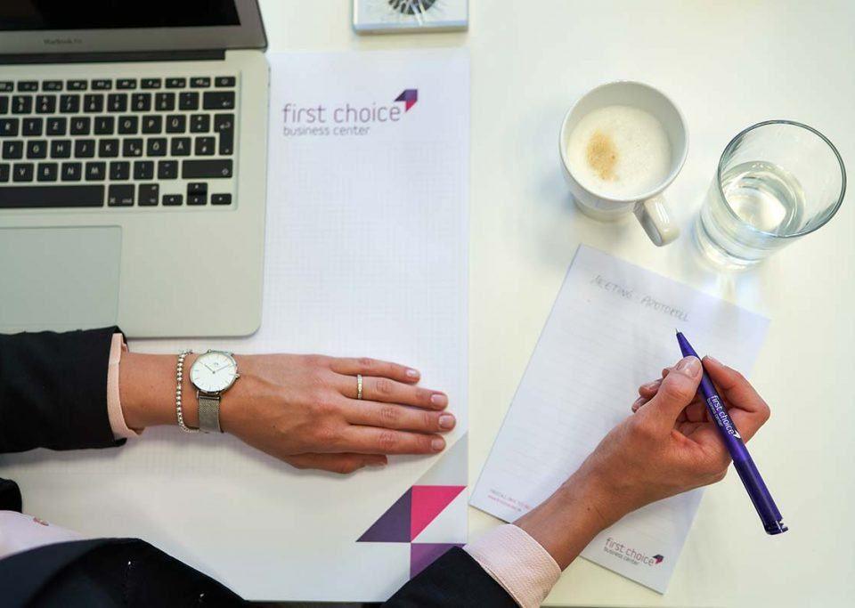 Auf einem weißen Schreibtisch steht ein silberner Laptop, daneben eine Kaffeetasse und ein Wasserglas sowie ein Schreibblock mit dem Logo der First Choice Business Center. Eine Person, von der man nur die Arme und Hände sieht, macht sich Notizen zum Thema Zeitmanagement auf dem Block.