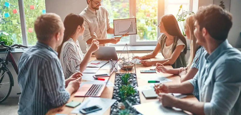 Was bedeutet ESG? Eine Gruppe junger, kreativer Menschen sitzt in einem lichtdurchfluteten Raum um einen langen Tisch und schaut auf eine Graphik, die ein junger Mann präsentiert